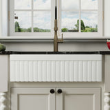 Front view of a white 33-inch reversible apron fireclay farmhouse sink installed beneath a brass faucet in a bright kitchen.