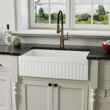NOZLOO NZ-3320T 33-inch reversible fireclay farmhouse sink installed fluted-side-out beneath a garden window with a black stone countertop and brass faucet.